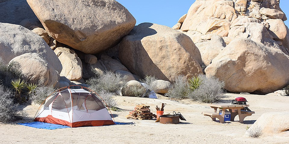 A test is pitched din the desert next to a pile of wood, a fire ring and a picnic table